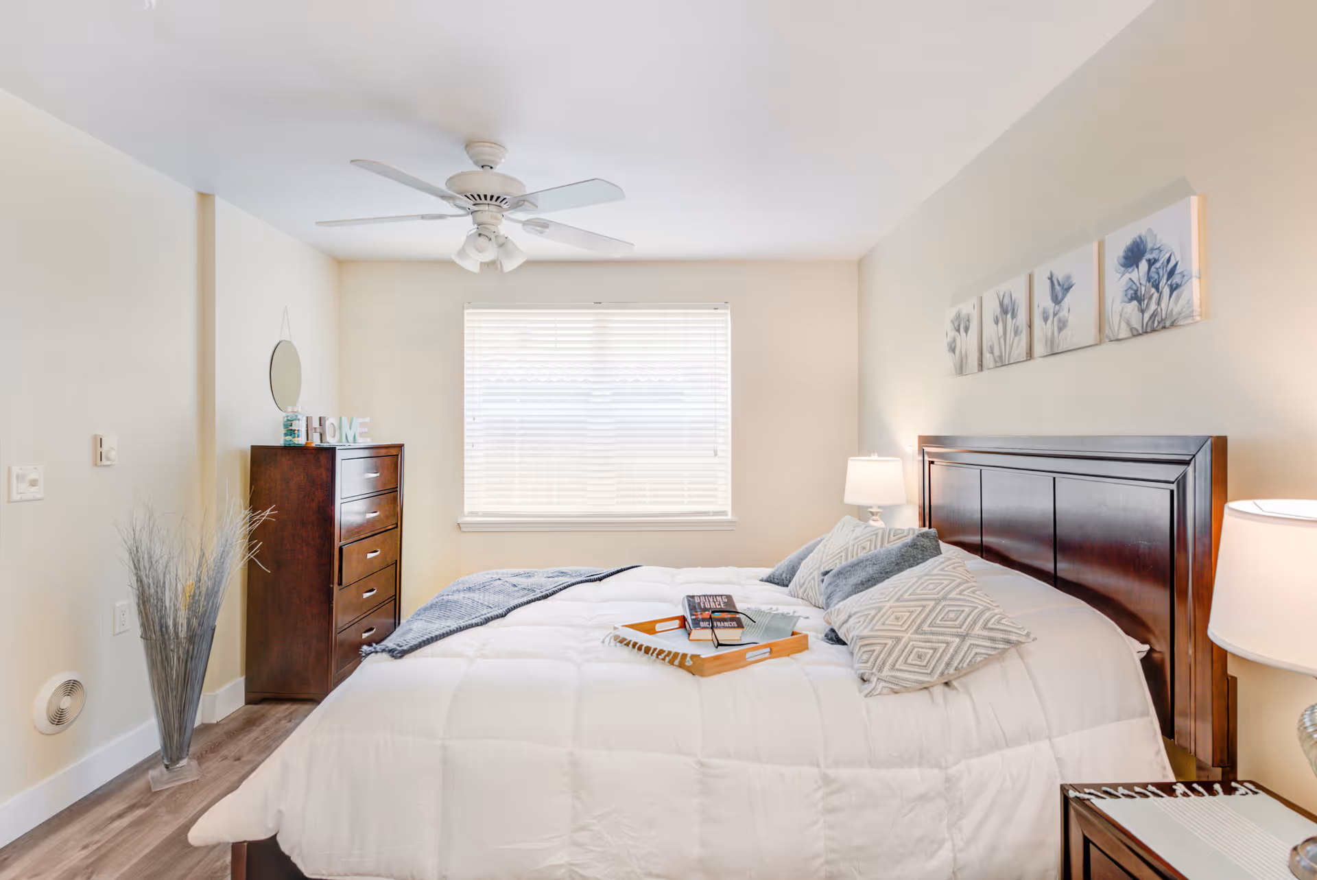 A bright bedroom with a large bed covered in a white comforter and multiple pillows. There is a wooden headboard and a wooden dresser with a round mirror and decorative items on top. A ceiling fan with lights is mounted on the ceiling, and a window with blinds lets in natural light. Two bedside lamps are on either side of the bed, and a wooden tray with books is placed on the bed.