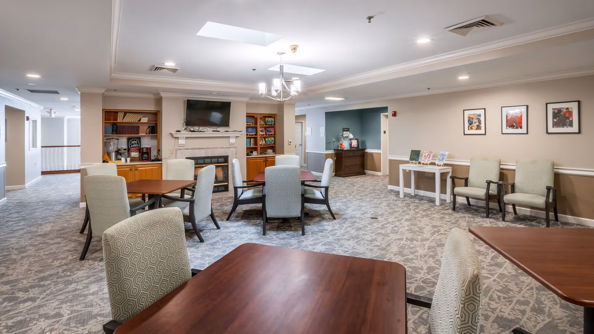 A spacious common area with multiple wooden tables and cushioned chairs arranged around the room. The room features a fireplace with a mounted flat-screen TV above it, built-in wooden shelves with books and decorative items, and a patterned carpet. The walls are adorned with framed pictures, and the ceiling has recessed lighting and skylights.