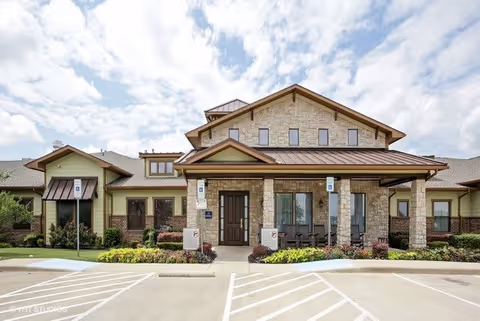 Front exterior view of Buffalo Creek Assisted Living and Memory Care facility showing a stone and wood building with a covered entrance, wheelchair accessible parking spaces, and landscaped greenery under a partly cloudy sky.