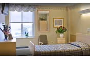 A tidy senior living bedroom with a single bed, chair, dresser, window, and floral arrangements.