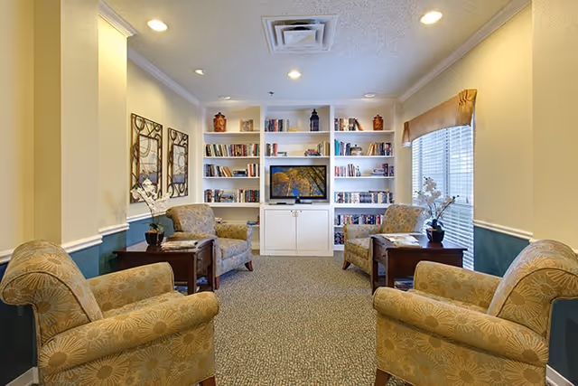 A cozy living room area with four patterned armchairs arranged around two wooden side tables. The room features built-in white bookshelves filled with books and decorative items, and a flat-screen TV in the center. There is a window with blinds and a valance on the right side, and two framed artworks on the left wall. The carpet is patterned, and the walls are painted in a light color with a teal wainscoting.