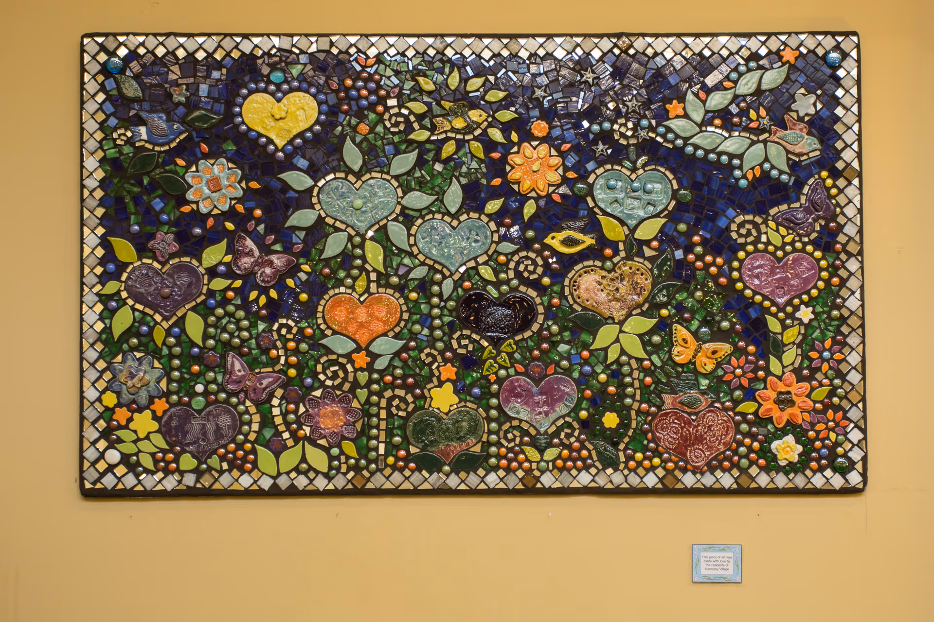 Colorful mosaic artwork mounted on a beige wall, featuring various hearts, flowers, butterflies, birds, and leaves in vibrant colors with a dark blue background and a white and black checkered border.
