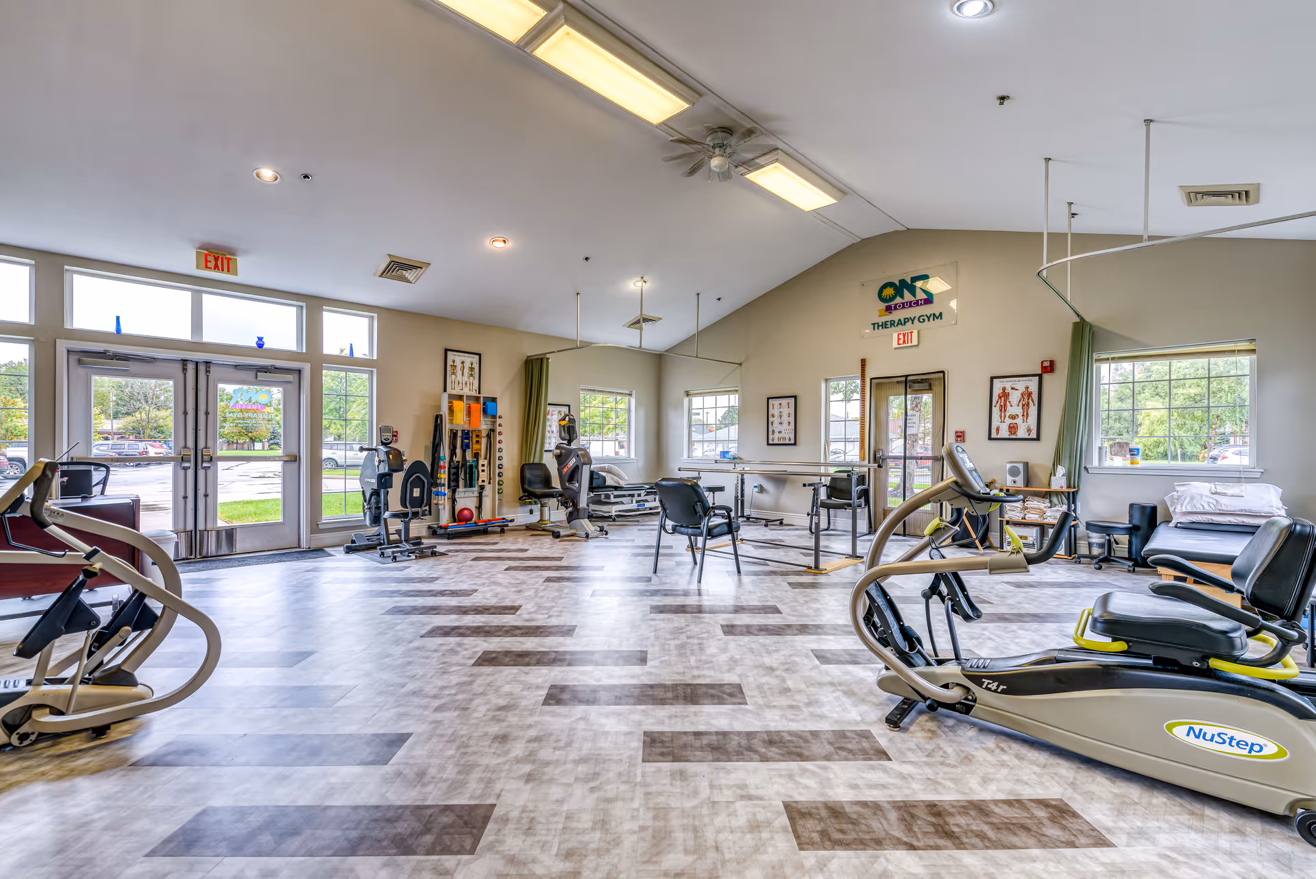 A spacious therapy gym with exercise equipment including NuStep machines, chairs, parallel bars, and various therapy tools. The room has large windows letting in natural light, light-colored walls, and a patterned floor. There are anatomical charts on the walls and exit doors visible at the back.
