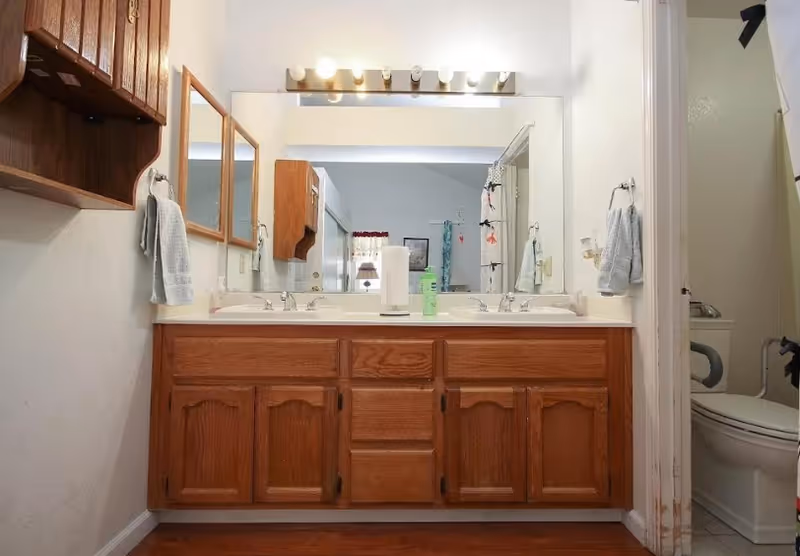 Double-sink bathroom vanity with mirror, cabinets, towels, and a toilet in a senior living home.