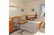 A small, tidy senior bedroom with a hospital-style bed, overbed table, armchair, and a small table by a curtained window.
