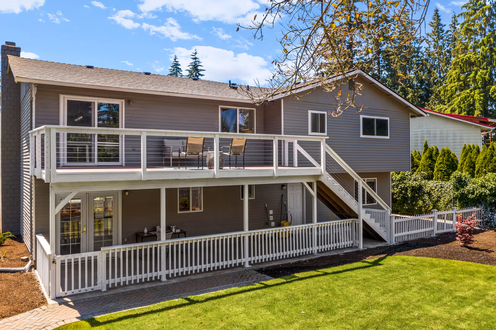 Exterior view of a two-story residential building with gray siding and white railings. The upper level features a balcony with two chairs and a small table, while the lower level has a covered patio area. There is a staircase on the right side leading from the ground to the balcony. The building is surrounded by a well-maintained lawn, trees, and shrubs under a partly cloudy blue sky.