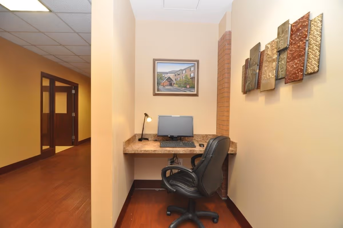 A small workspace nook with a computer monitor, keyboard, mouse, and desk lamp on a marble-patterned countertop. A black office chair is positioned in front of the desk. The walls are beige with a decorative metal wall art on the right and a framed picture above the desk. The floor is wooden, and a hallway with wooden doors is visible to the left.