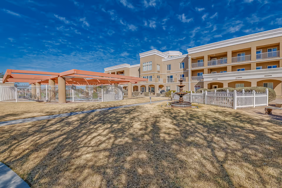 Front exterior of a senior living facility with a three-story beige building, covered patio, fountain, white picket fence and grassy courtyard under a blue sky.