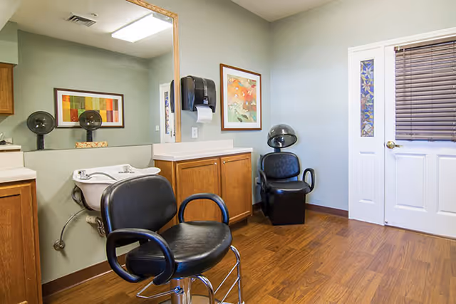 Small salon-style interior with two black styling chairs, a shampoo sink, large mirror, wooden cabinets, and a door.