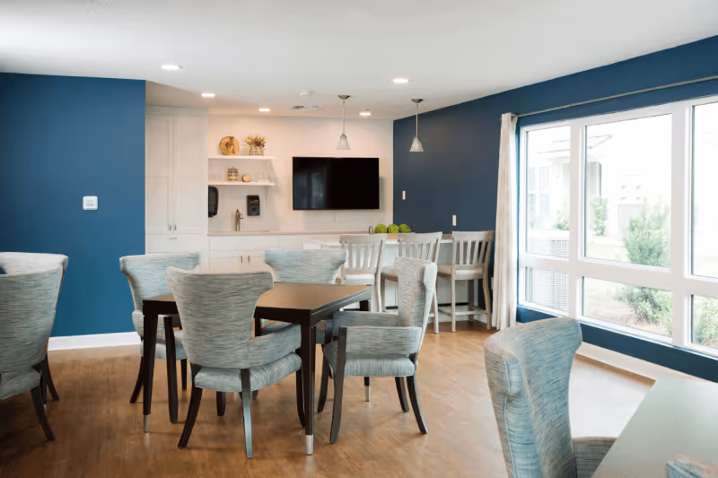 A bright and modern common area with several tables and cushioned chairs arranged on a wooden floor. The walls are painted blue and white, with a large window letting in natural light. There is a flat-screen TV mounted on the wall above a white cabinet, and a counter with bar stools in the background.