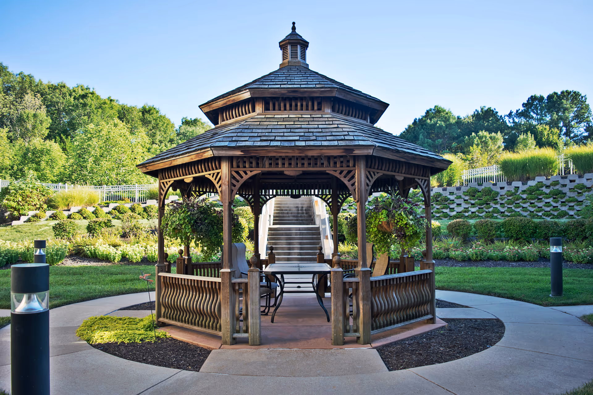 A wooden gazebo with a shingled roof situated in a landscaped garden area with green grass, plants, and trees in the background. Inside the gazebo, there is a table and chairs. A paved walkway surrounds the gazebo, and there are modern outdoor lights along the path.