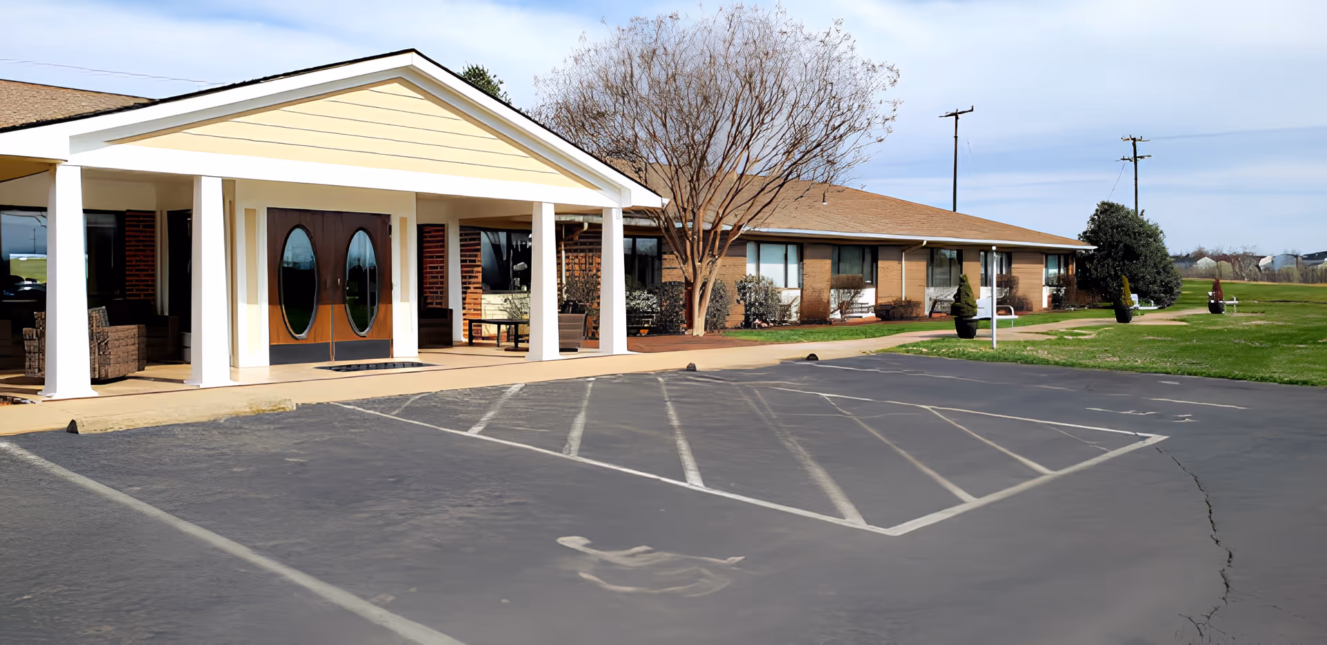 Exterior view of Blue Ridge Christian Homes facility showing a single-story building with a covered entrance supported by white columns, double wooden doors with oval windows, a parking lot with a handicapped parking space, and a grassy area with trees and shrubs.