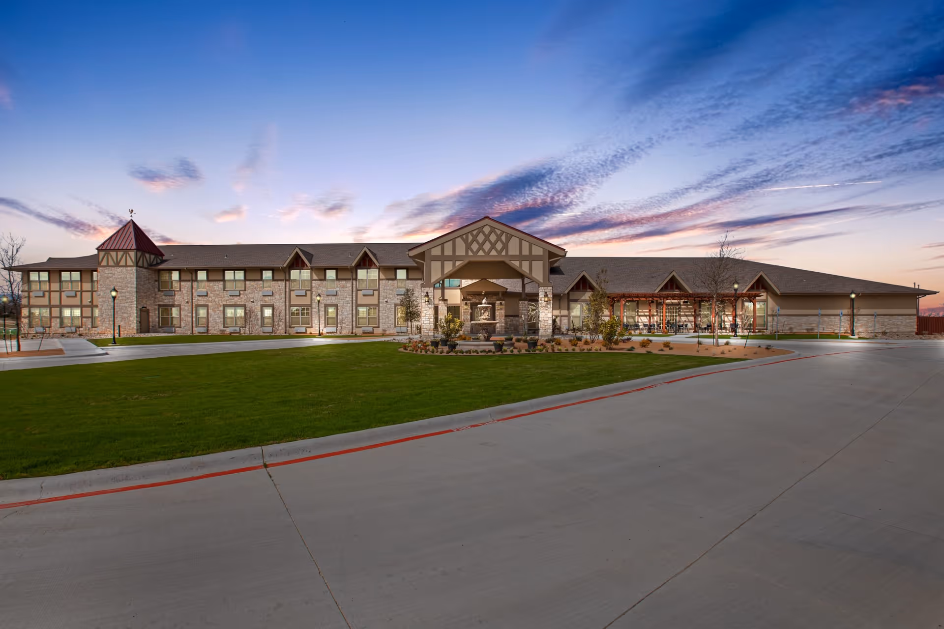 Exterior view of The Villages of Windcrest senior living facility at dusk, showing a two-story building with stone and beige siding, a covered entrance with a fountain, well-maintained green lawn, and a paved driveway.