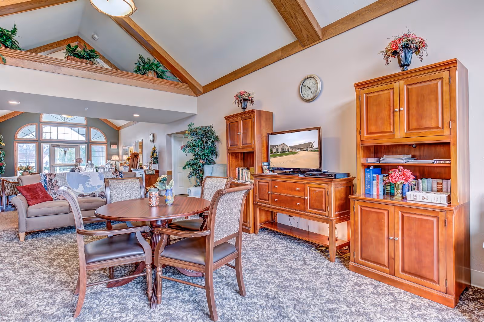A cozy living room area in a senior living facility with a round wooden table and four chairs in the foreground. Behind the table, there are two wooden cabinets with decorative flowers on top, a flat-screen TV, and a clock on the wall. The room has a high ceiling with wooden beams and plants placed on a ledge near the ceiling. In the background, there is a seating area with armchairs and a couch near large windows letting in natural light.