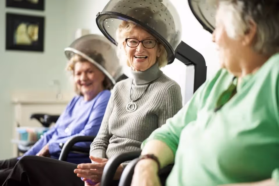 Three elderly women sitting under hair dryers in a salon, smiling and engaging in conversation. The woman in the center wears glasses and a gray sweater, while the others wear purple and green tops.