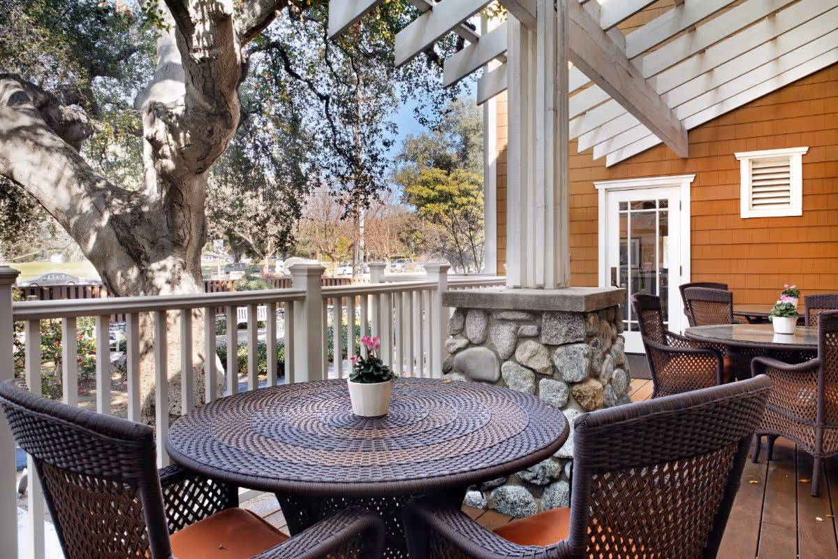 Outdoor covered patio with round wicker tables and chairs on a deck overlooking trees and a garden.