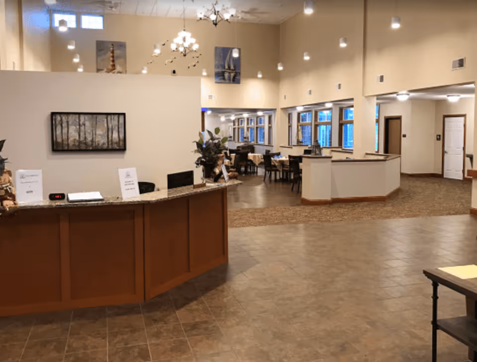Interior view of a senior living facility lobby with a curved wooden reception desk in the foreground. Behind the desk, there is a large open area with tables and chairs arranged for dining or socializing. The walls are decorated with framed artwork, and multiple ceiling lights illuminate the space. Large windows line the far wall, allowing natural light to enter.