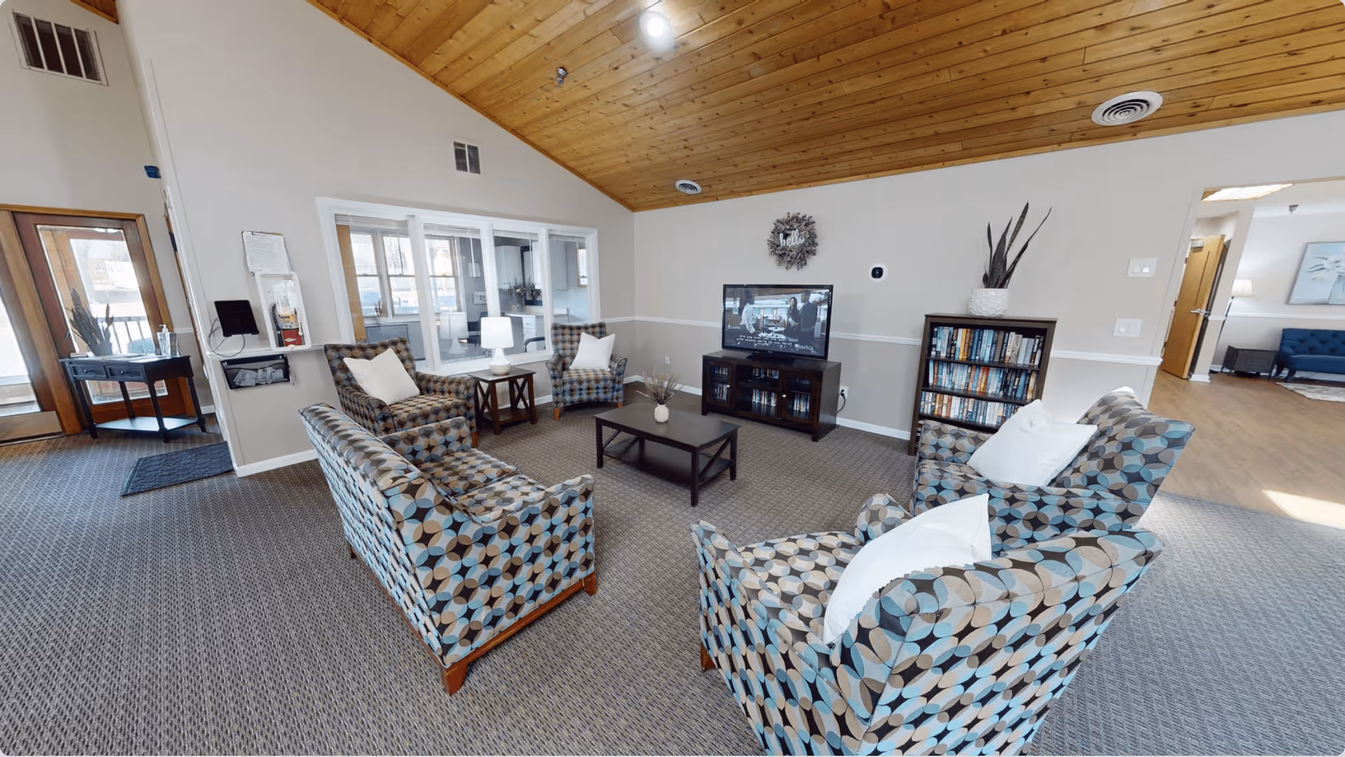 A cozy living room in Aspen Assisted Living featuring patterned armchairs and a sofa arranged around a dark wood coffee table. A TV is placed on a stand against the wall, with a bookshelf nearby holding various books. The room has a carpeted floor, a wooden ceiling, and neutral-colored walls with a decorative wreath above the TV. A window looks into an adjacent room, and a door leads outside.