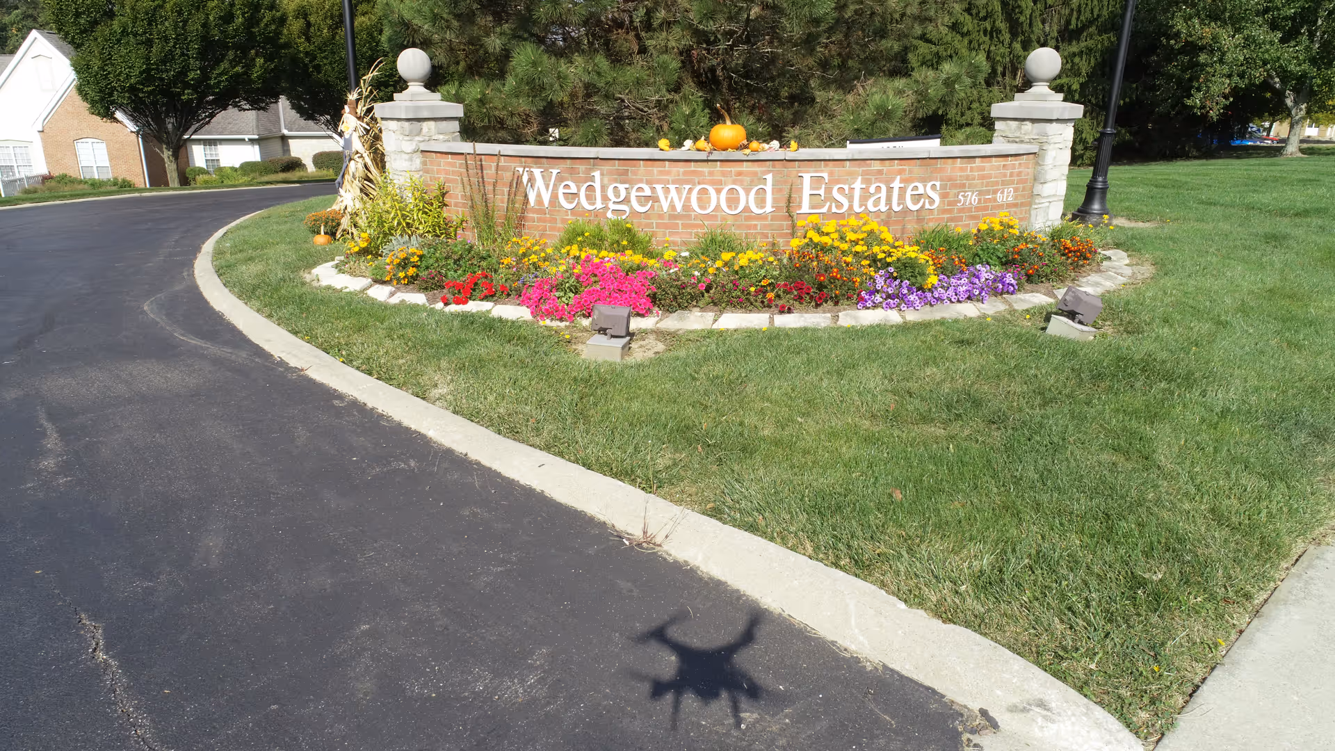 Entrance sign for Wedgewood Estates made of brick with white lettering, surrounded by colorful flowers and green grass, with a paved road curving around it and houses and trees in the background.