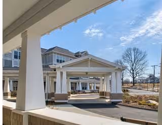 Exterior view of a senior living facility showing a covered entrance with white pillars and a driveway. The building has multiple windows and a beige exterior. There is a clear blue sky with some clouds and a leafless tree in the background.
