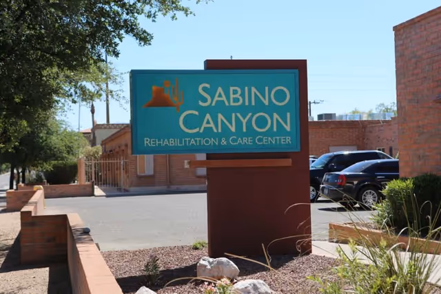 Outdoor view of the Sabino Canyon Rehabilitation & Care Center sign with a desert landscape design, surrounded by brick walls, plants, and parked cars in the background under a clear sky.