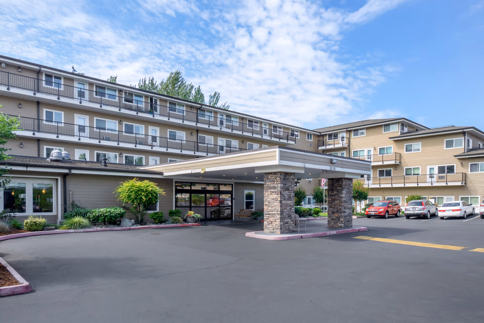 Exterior view of a multi-story senior living facility with a covered entrance supported by stone pillars, several parked cars, landscaped greenery, and a partly cloudy sky.