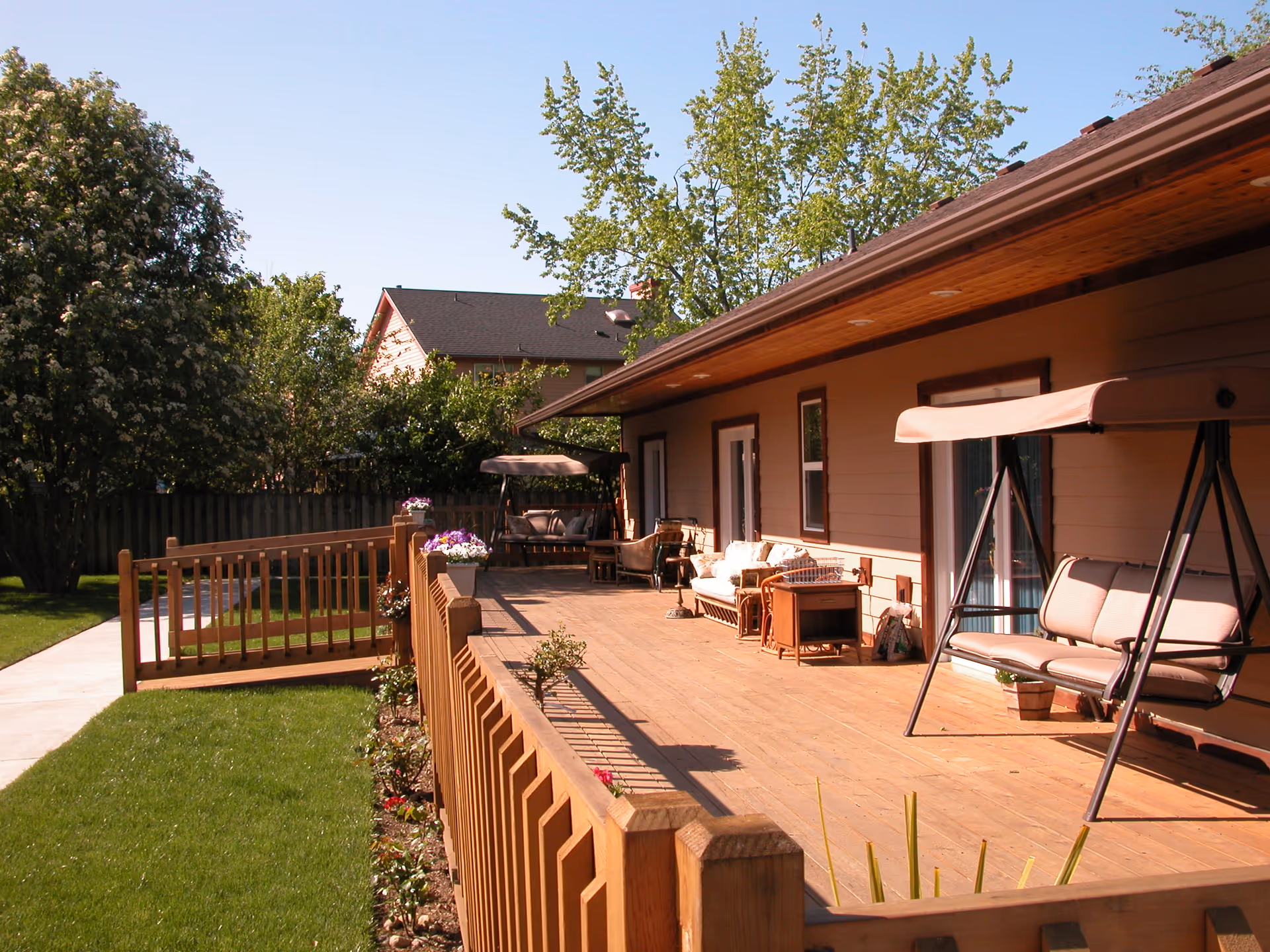 A spacious wooden deck attached to a single-story building with outdoor seating including cushioned swings and chairs. The deck is surrounded by a wooden fence and overlooks a well-maintained lawn with trees and flower beds. The sky is clear and blue.