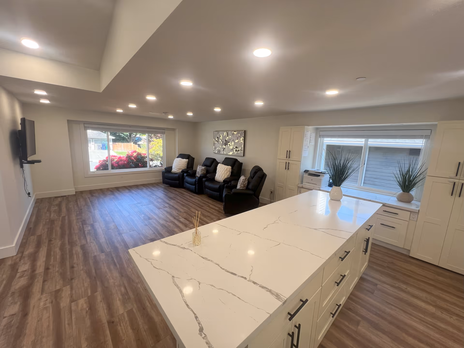 A bright and spacious common area in a senior care facility featuring a large white marble countertop island with drawers, three black reclining chairs with cushions against the far wall, a wall-mounted TV, large windows letting in natural light, and two potted plants on the countertop near the window.