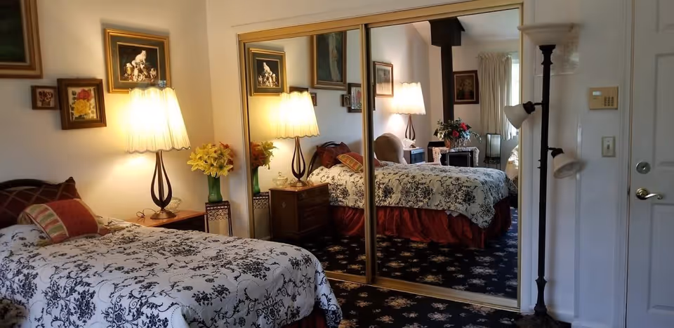 A cozy bedroom with a single bed covered in a white and black floral bedspread and a red bed skirt. There is a bedside table with a lamp and a vase of yellow flowers. The wall behind the bed has several framed pictures. A large mirrored closet door reflects the room, showing another bed and additional furniture. A tall floor lamp stands near a white door with a gold handle.