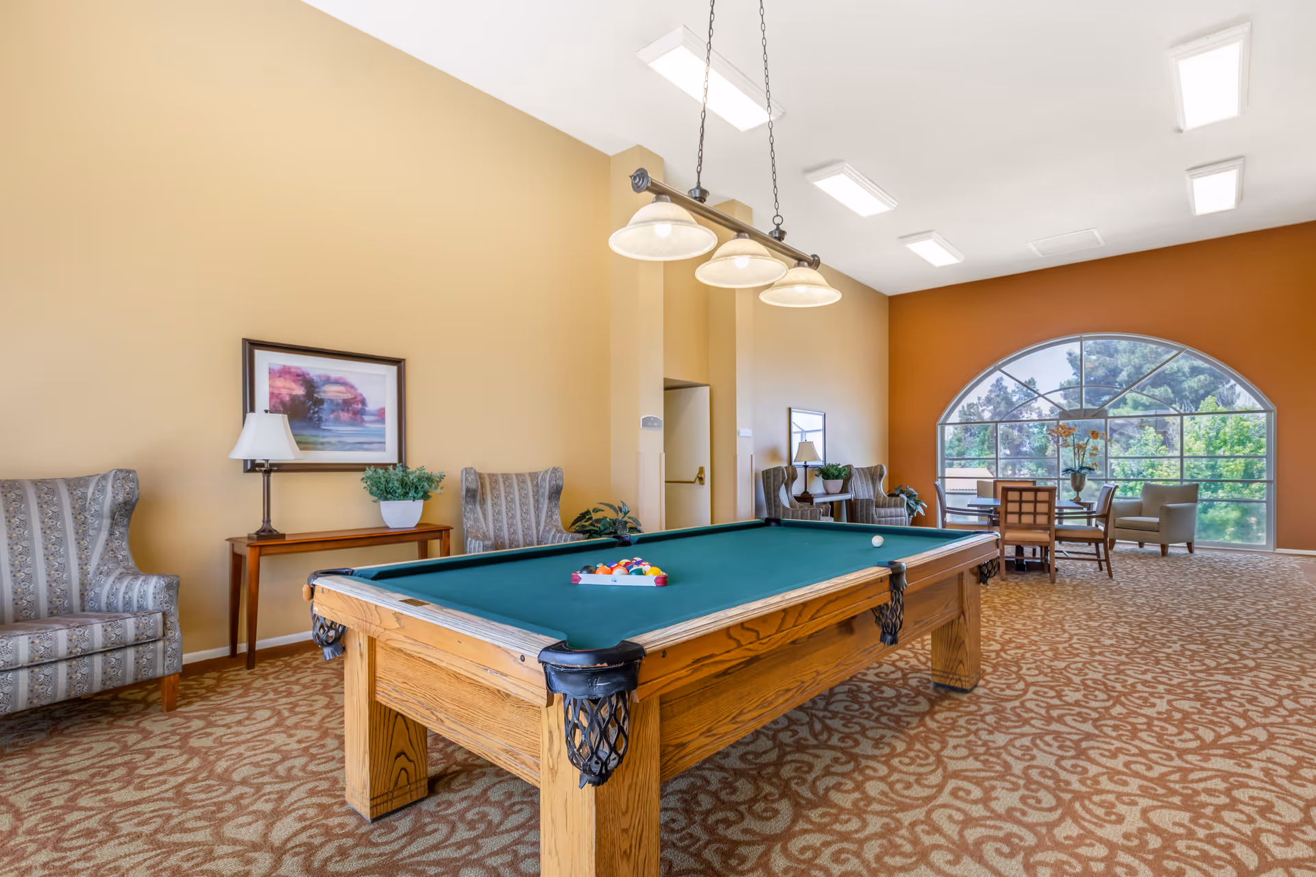 A spacious recreational room with a pool table in the center, surrounded by patterned armchairs and a wooden table with a lamp and plant. The room has beige and orange walls, a large arched window letting in natural light, and a carpet with a swirling pattern.
