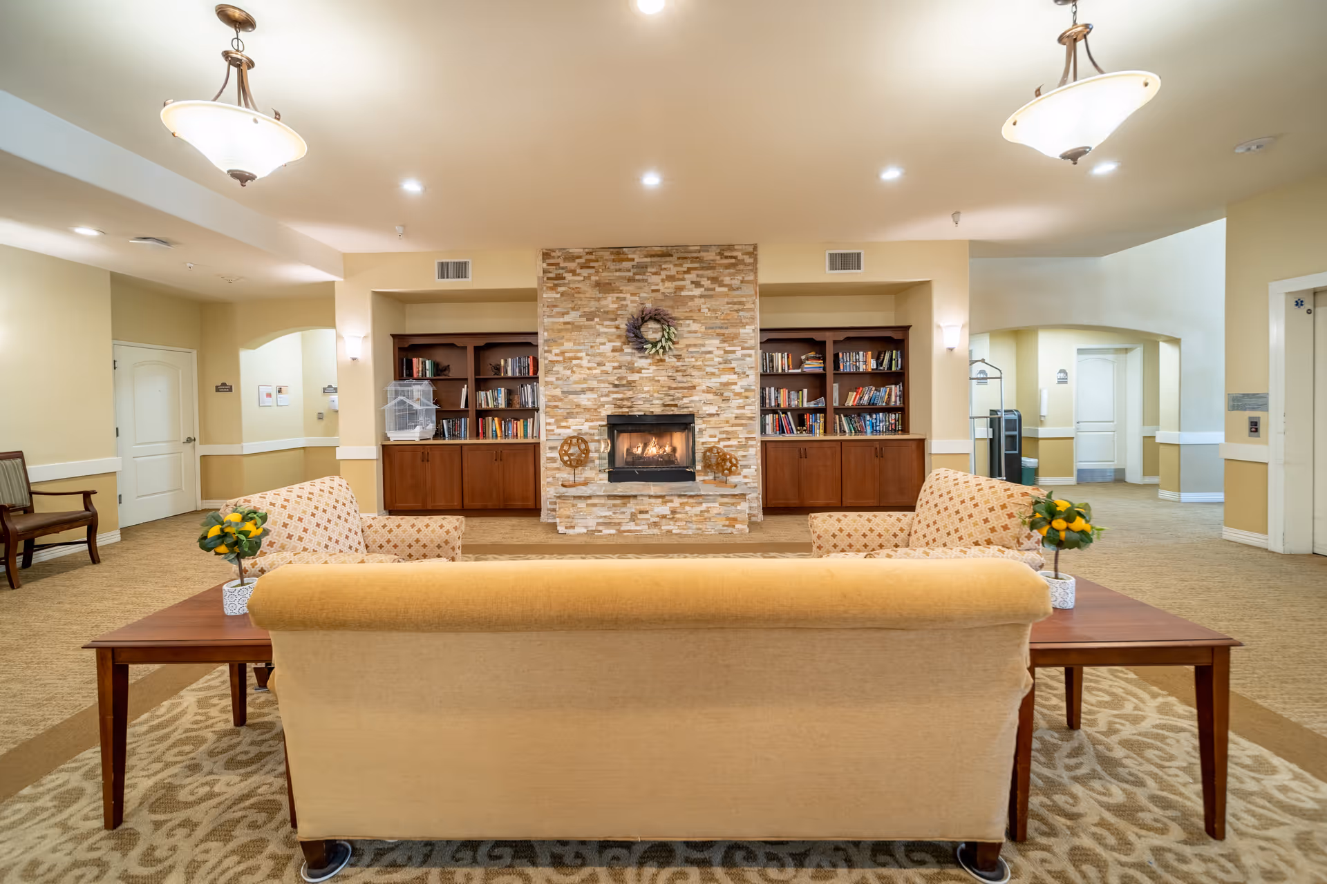 A cozy living room area in a senior living facility featuring a beige sofa and two patterned armchairs arranged around a stone fireplace with a wreath above it. On either side of the fireplace are wooden bookshelves filled with books. The room is well-lit with ceiling lights and two hanging light fixtures. There are small tables with potted plants next to the sofa, and doorways and hallways are visible in the background.