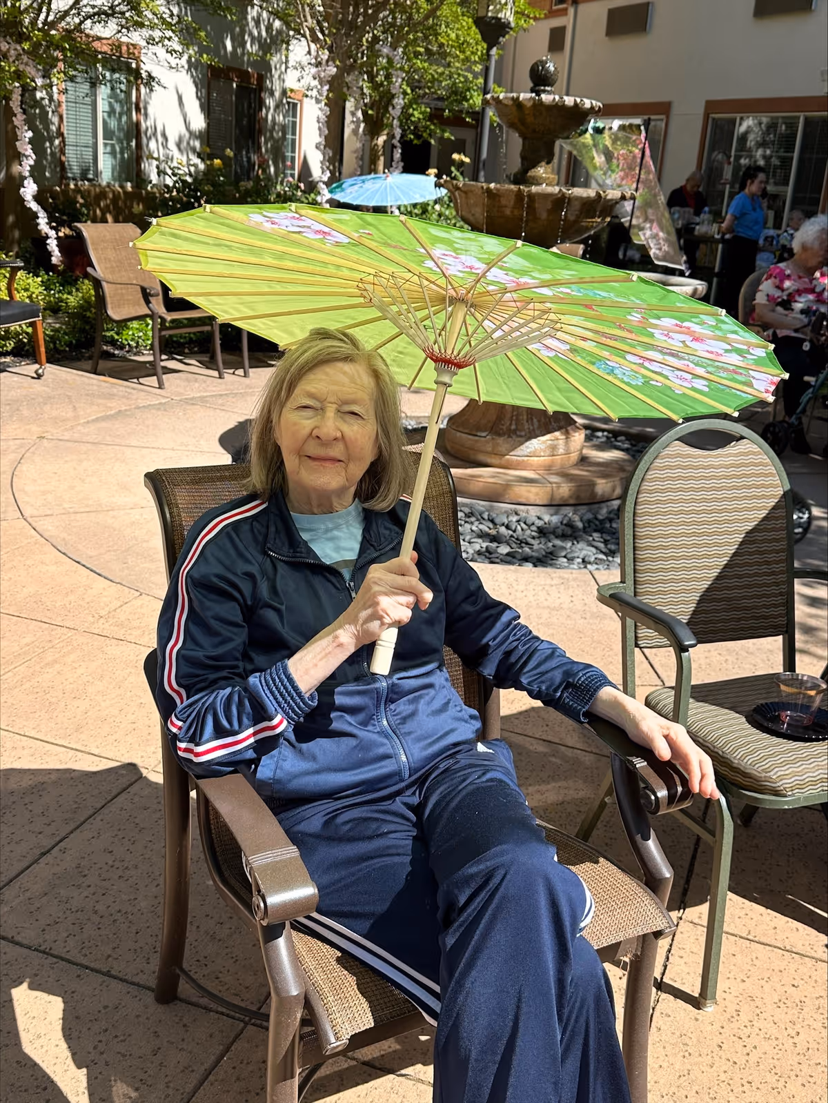 An elderly woman sitting outdoors on a patio chair holding a green floral parasol above her head. She is wearing a navy blue tracksuit and is seated near a stone fountain with other chairs and people visible in the background.
