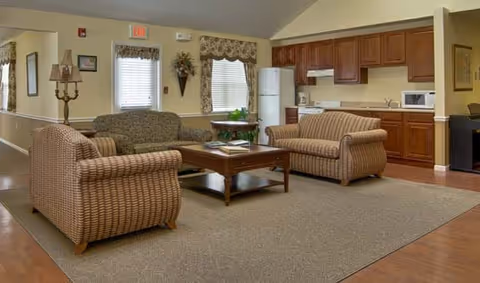 A cozy living room area with two patterned sofas and one armchair arranged around a wooden coffee table. Behind the seating area is a kitchenette with wooden cabinets, a white refrigerator, microwave, and countertop. The room has beige walls, windows with valances, and a carpeted floor with hardwood around the edges.