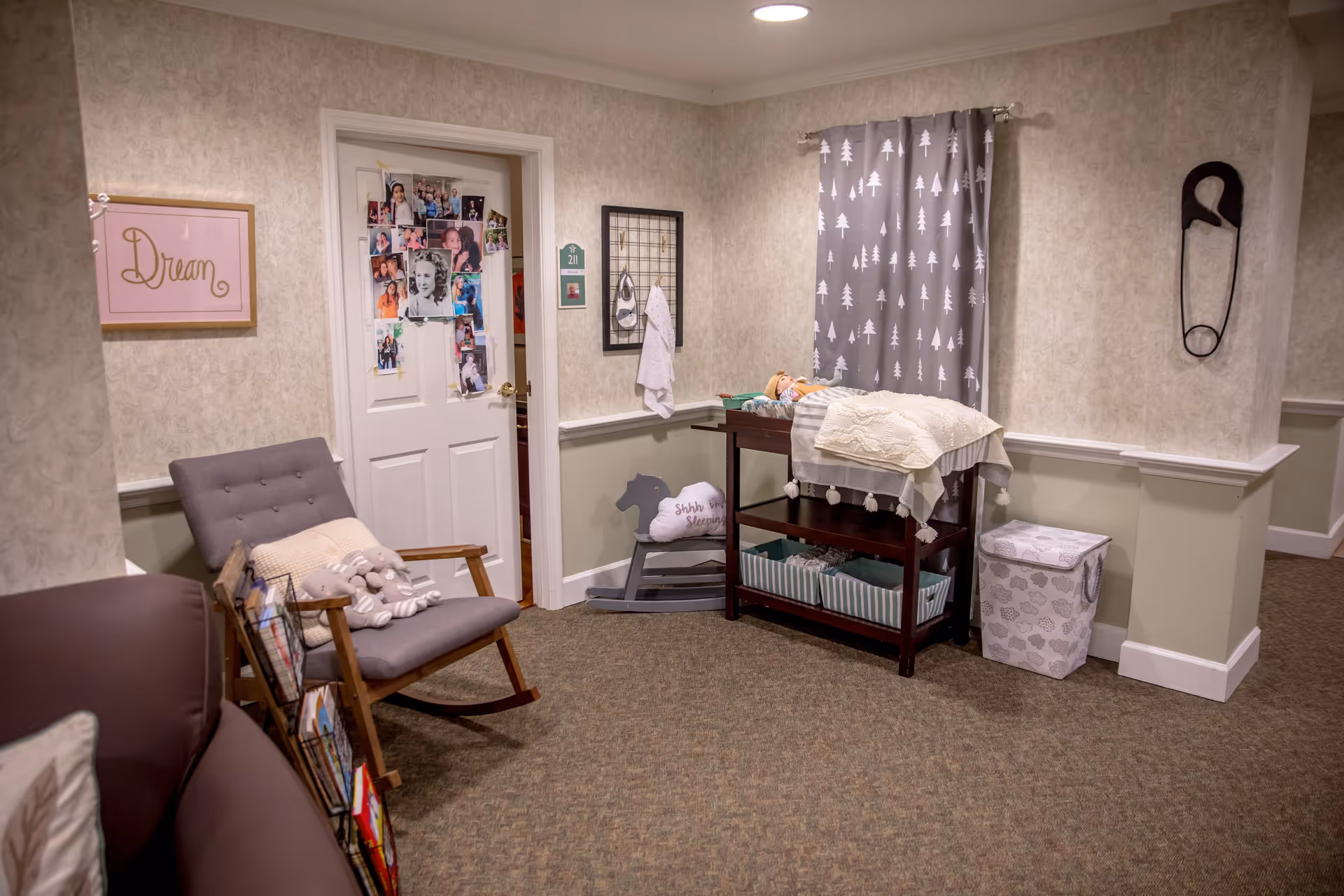 A cozy corner in an assisted living facility hallway featuring a gray rocking chair with a stuffed animal and pillow, a changing table with a doll on top, a rocking horse with a pillow that says 'Shhh... I'm Sleeping,' a laundry hamper with cloud patterns, and a door decorated with multiple family photos. The walls are light-colored with a framed 'Dream' sign and a large safety pin decoration.