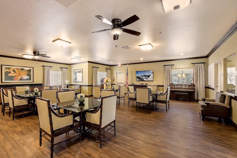 A spacious and well-lit common dining area with multiple tables and chairs arranged neatly. The room features wooden flooring, ceiling fans, framed artwork on the walls, a piano near the window, and a jukebox in the corner. Large windows with curtains allow natural light to enter the room.