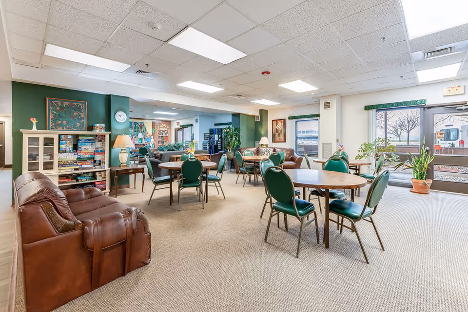 A spacious common area in a senior living facility with several round tables surrounded by green chairs, a brown leather couch, a cabinet filled with board games, potted plants near large windows and glass doors, and soft overhead lighting.