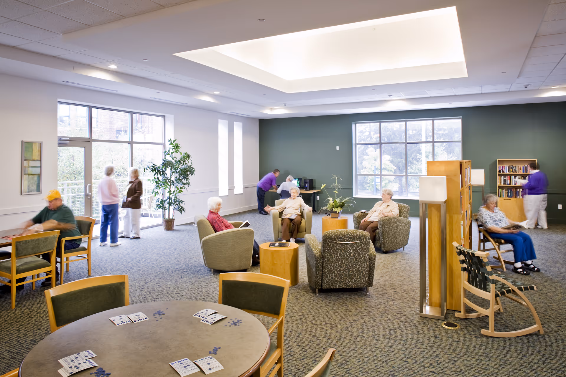 A spacious, well-lit common area in Woodcrest Retirement Residence with several elderly people seated in comfortable chairs and engaging in activities. The room features large windows, a skylight, bookshelves, and tables with chairs. Some people are playing games, reading, or standing near the windows.