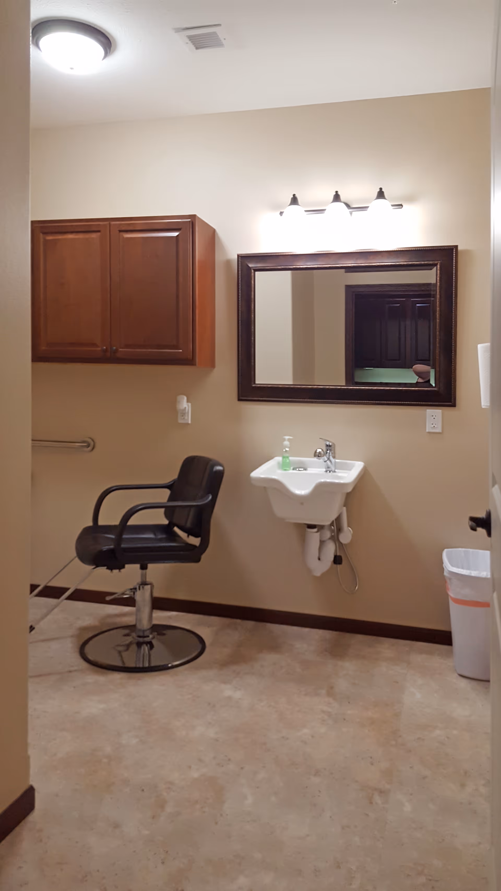 Interior view of a room with a black salon chair, a wall-mounted sink with a soap dispenser, a large framed mirror above the sink, a wooden cabinet mounted on the wall, and a trash can in the corner. The room has beige walls and a tiled floor.