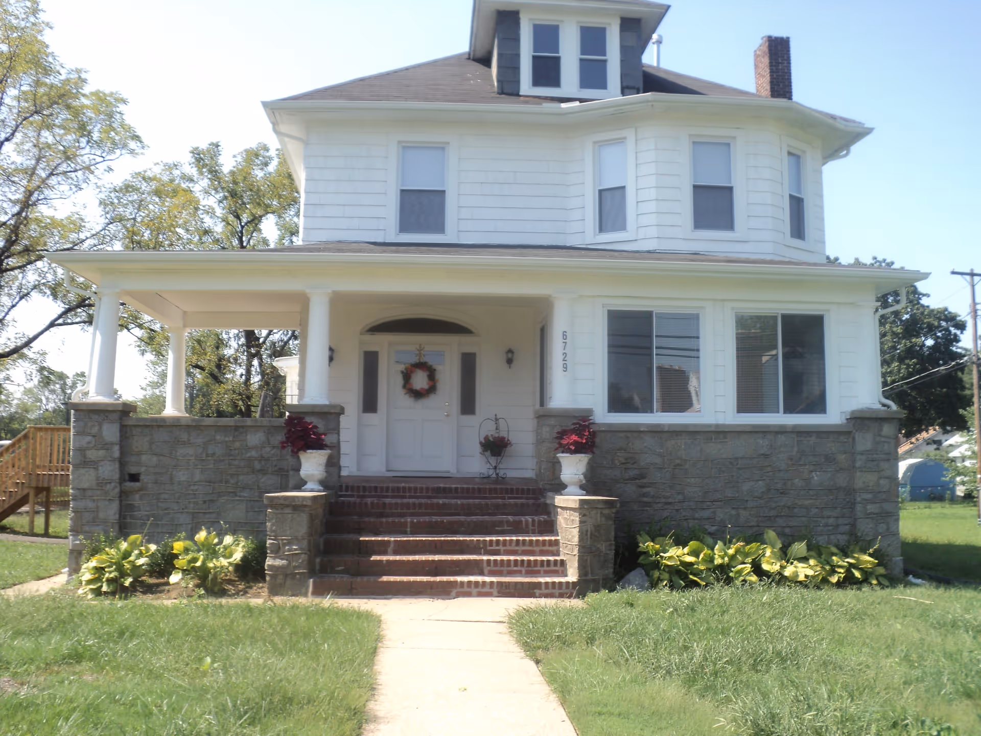 Front exterior view of a two-story white house with a stone foundation, a covered porch with columns, a brick staircase leading to the front door decorated with a wreath, and surrounding greenery.