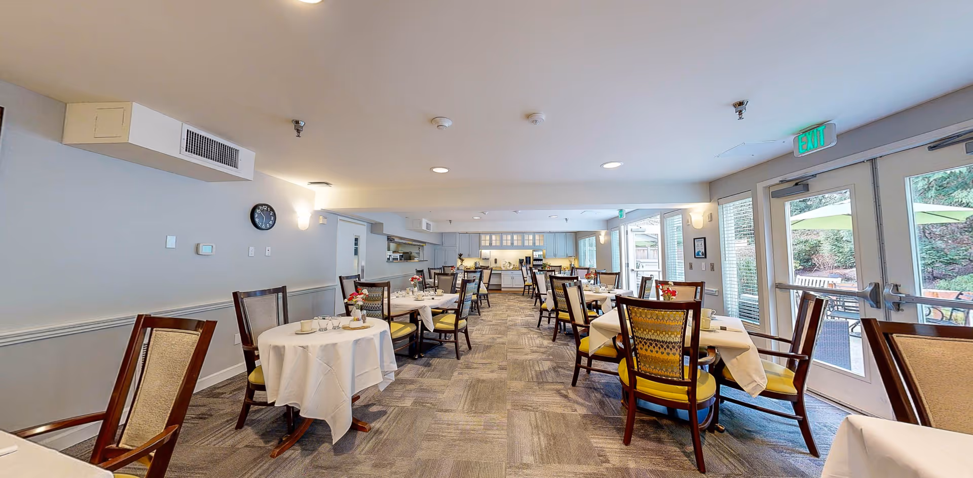 Bright assisted-living dining room with round tables set with white tablecloths, chairs, and glass doors leading to an outdoor patio.