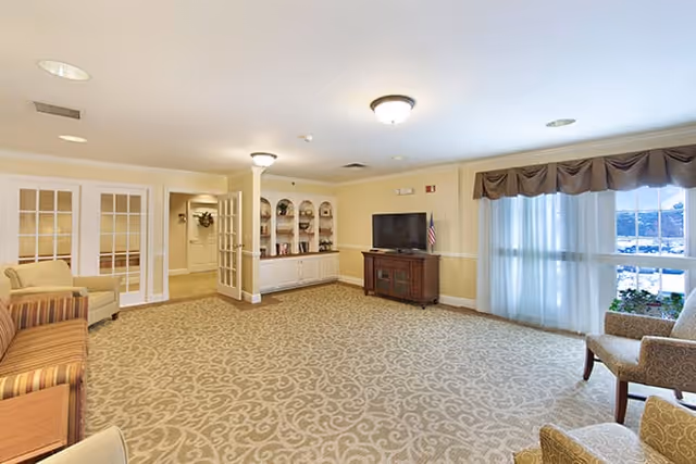 A spacious living room area in a senior living facility with beige patterned carpet, cream-colored walls, and ceiling lights. The room features a striped sofa, armchairs, a wooden TV stand with a flat-screen television, built-in shelves with decorative items, and large windows with sheer curtains and valances letting in natural light.