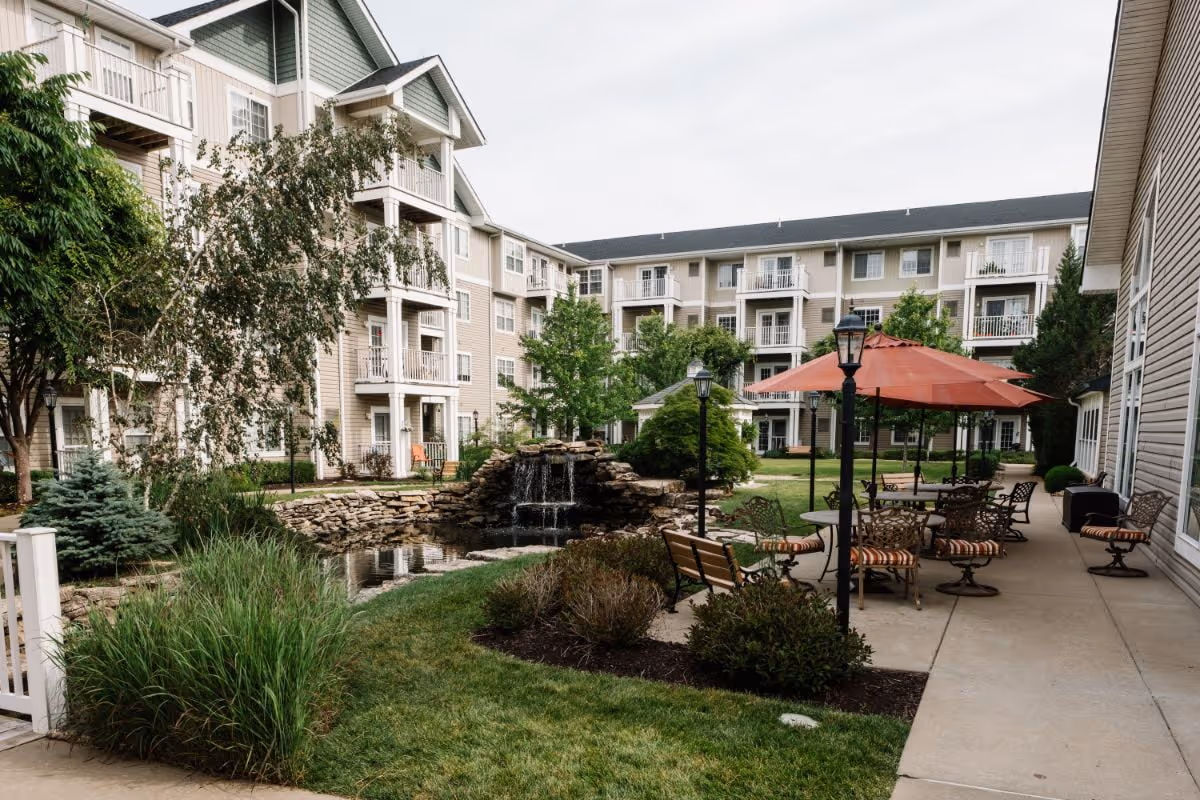 Outdoor courtyard area of a senior living facility with a small waterfall feature, pond, green grass, shrubs, and trees. There are several patio tables with chairs and large red umbrellas on a concrete walkway. The background shows a multi-story beige building with balconies and white trim.