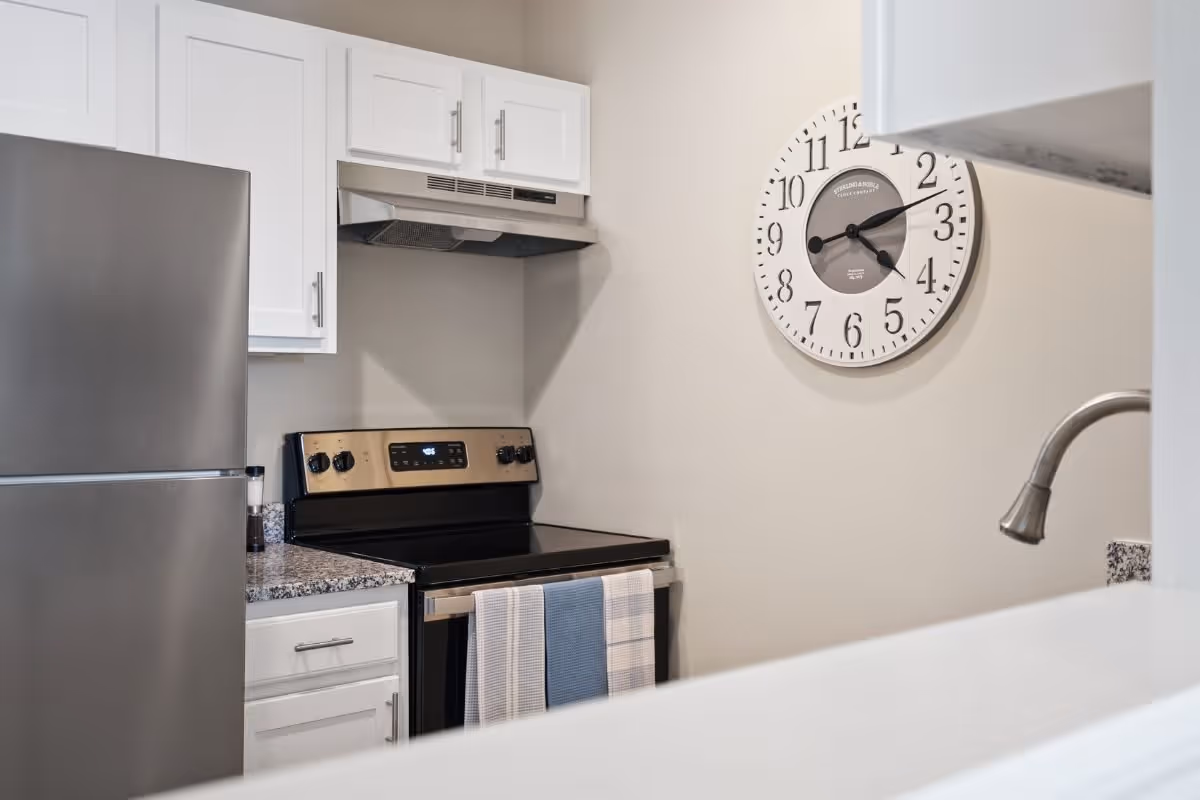 A modern kitchen area featuring a stainless steel refrigerator, an electric stove with a range hood, white cabinets, a granite countertop, and a wall clock showing the time as 4:15. A faucet and part of a sink are visible in the foreground.