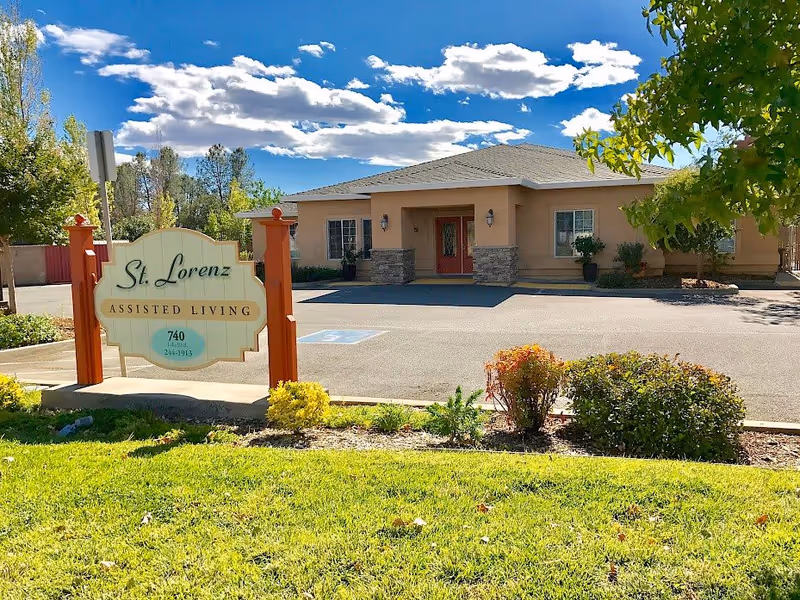 Exterior view of St Lorenz Assisted Living facility on a sunny day with a clear blue sky and scattered clouds. The building is a single-story structure with beige walls, a gray roof, and a red front door. In front of the building is a sign that reads 'St. Lorenz Assisted Living' with the address and phone number. There are small bushes and green grass in the foreground.