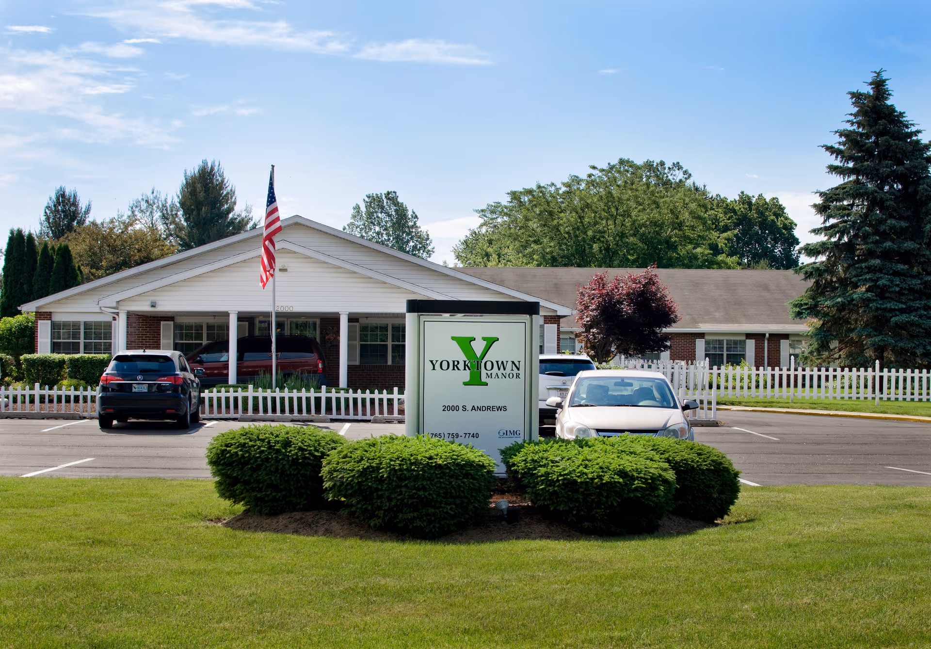 Exterior view of Yorktown Manor, a single-story brick and white building with a pitched roof, surrounded by green bushes and trees. There is a parking lot with several cars and an American flag on a flagpole in front of the building. A sign in front displays the name Yorktown Manor and the address 2000 S. Andrews.