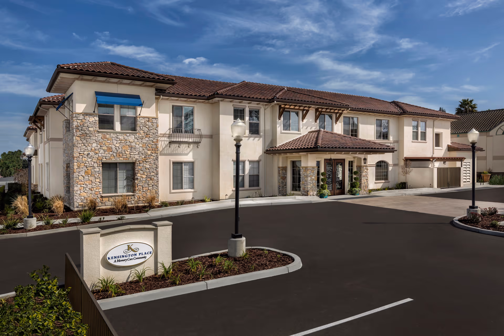 Exterior view of Kensington Place Redwood City, a two-story building with stone and stucco facade, tiled roof, and a driveway with street lamps. There is a sign in front that reads 'Kensington Place A Memory Care Community'.