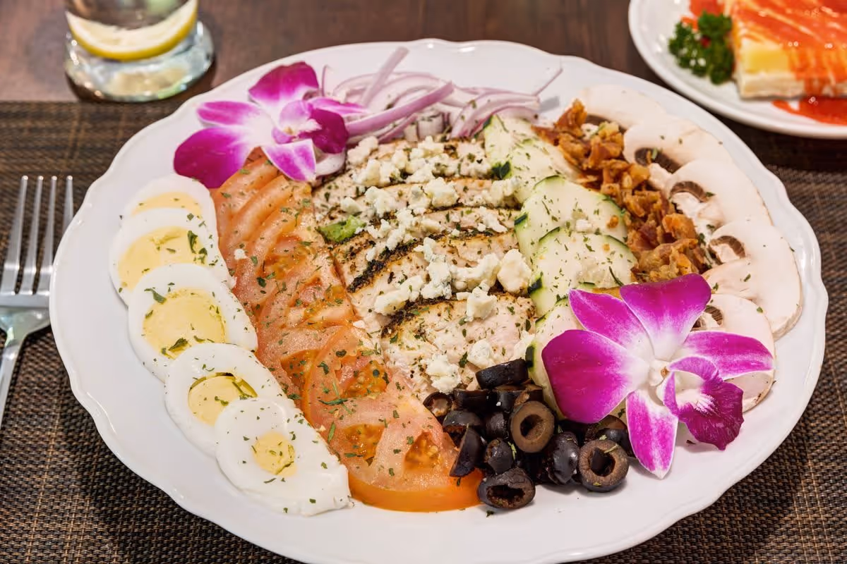A white plate with a colorful salad arranged in sections including sliced hard-boiled eggs, tomato slices, grilled chicken topped with crumbled cheese, cucumber slices, black olives, mushrooms, red onion slices, and crispy bacon bits, garnished with two purple orchid flowers. A fork is placed to the left of the plate and a glass of water with a lemon slice is partially visible in the background.