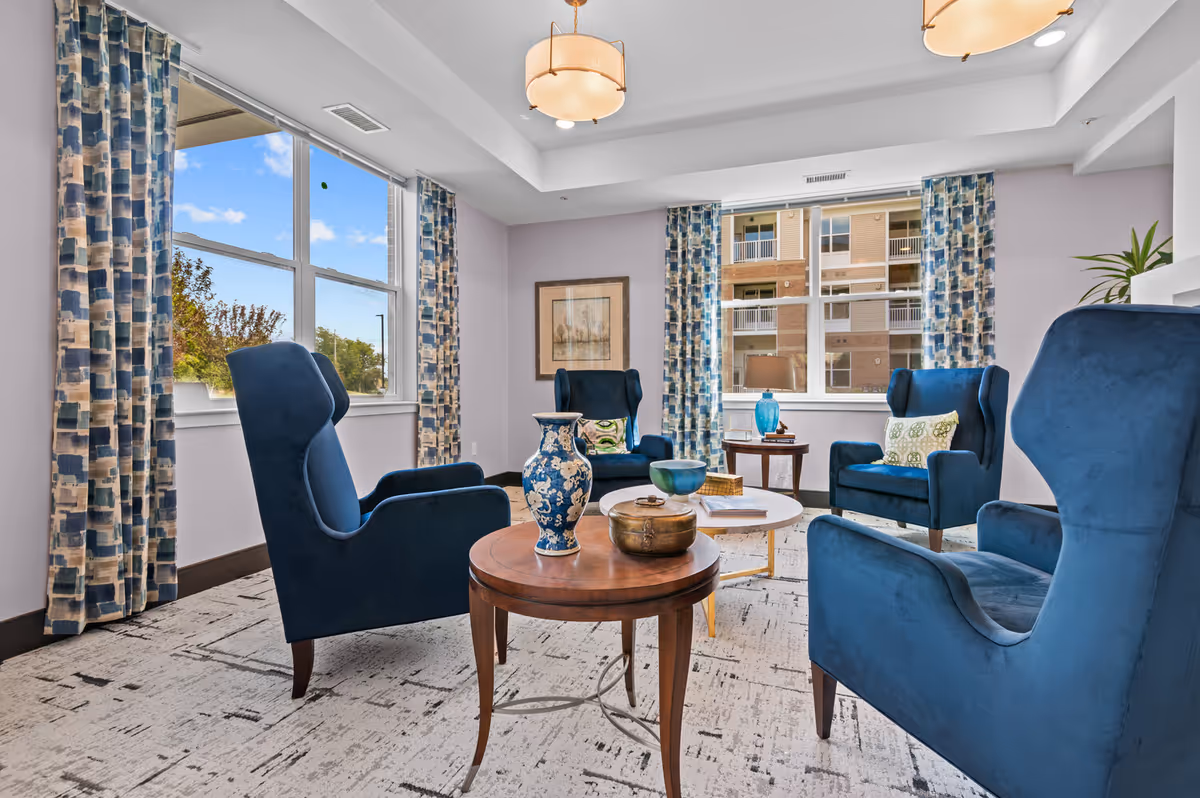 A bright and cozy sitting area with four blue upholstered wingback chairs arranged around two round wooden tables and a white coffee table. The tables hold decorative items including a blue and white vase, a brass container, a blue bowl, and books. The room has large windows with patterned blue and beige curtains, allowing natural light to fill the space. A framed artwork hangs on the wall, and a blue lamp sits on a side table near one of the windows. The carpet has a modern abstract pattern in neutral tones.