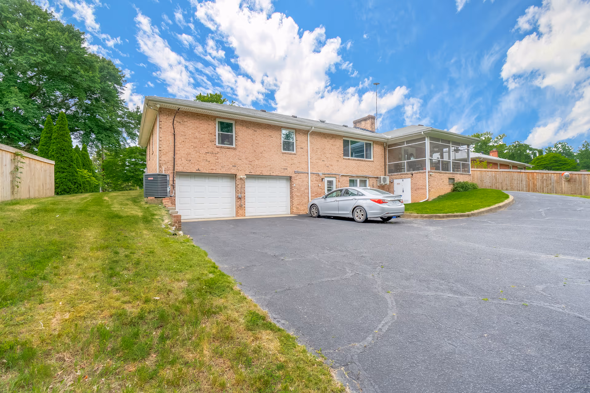 Exterior view of a two-story brick building with two white garage doors, a silver car parked in front, a fenced yard with green grass and trees, and a partly cloudy blue sky.