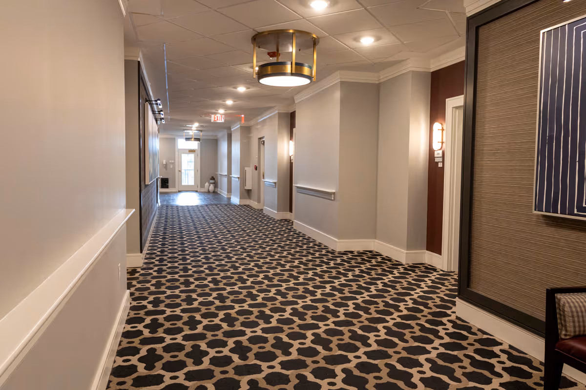 A well-lit hallway in a senior living facility with patterned carpet, beige walls, ceiling lights, and a door at the far end. There are handrails along the walls and some decorative elements including a framed artwork and a chair partially visible on the right.